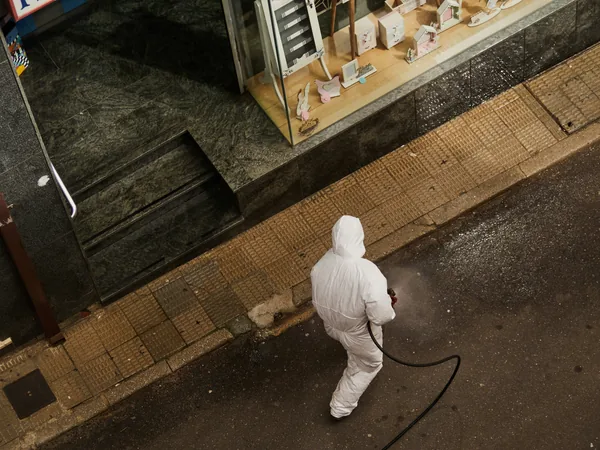 worker in protective suit spraying storefront pavement with cleaning hose